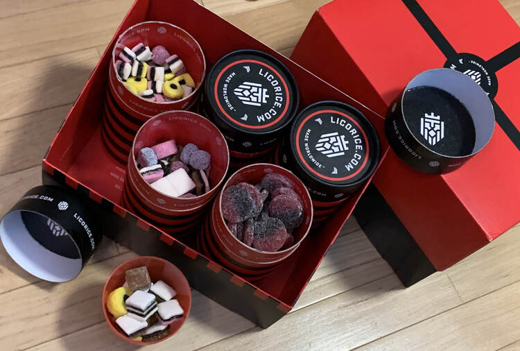 Image shows a closed gift box of licorice to the right and an opened box to the left. At the bottom left there's a red plastic cup with licorice in it next to several opened canisters. 