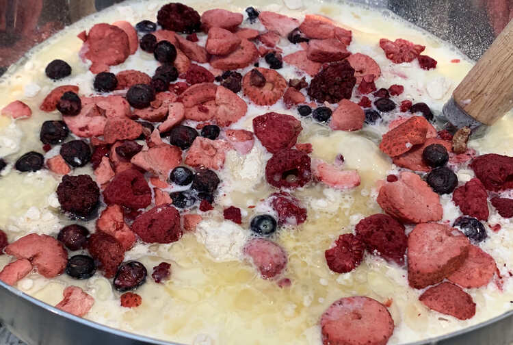 Image shows a closeup shot of a metal bowl with white greasy mixture topped with dried out strawberries, blueberries, the odd blackberry, and the handle of a whisk. 