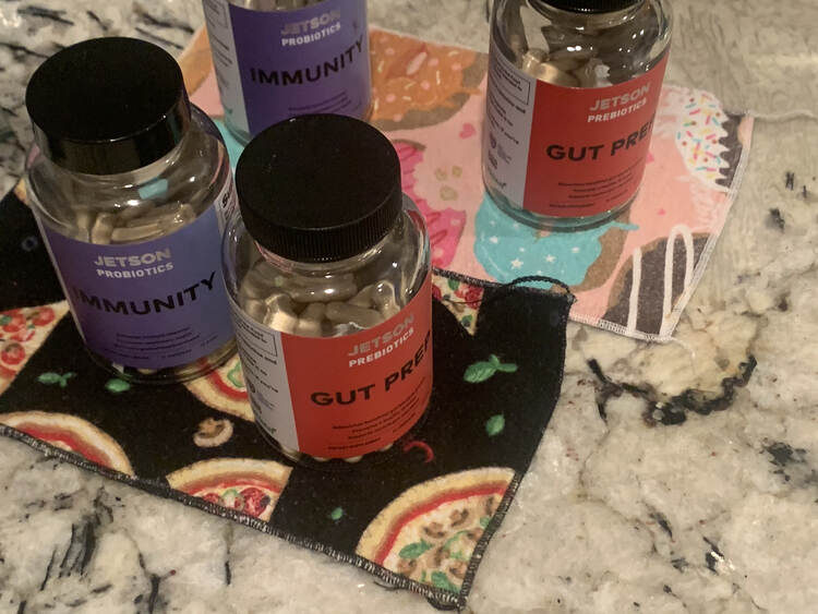 Image shows two purple "immunity" bottles and two "gut prep" bottles. In the fore front one of each rests on a black pizza adorned cloth napkins while in the background the other two bottles rest on a pink donut festooned napkin. 