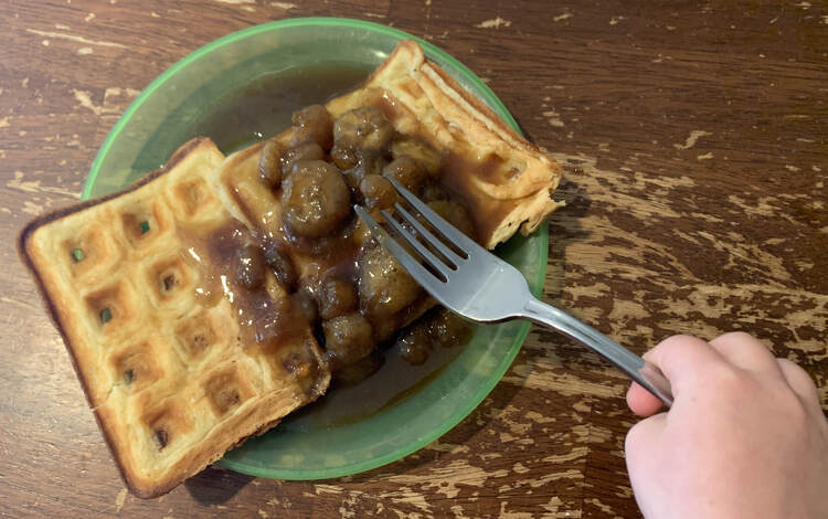 Image shows two waffles side by side on a green plastic plate covered in brown lumps with brown sauce around it. A hand holding a fork is pressed on the top. 