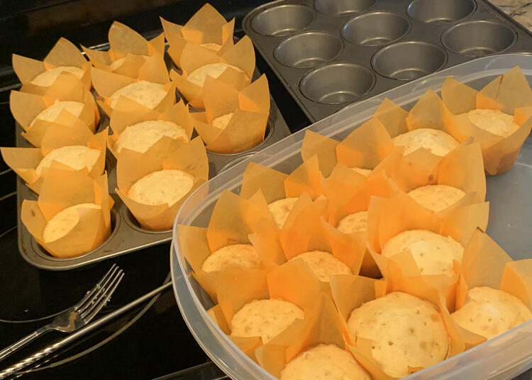 Image shows the baked cupcakes after they had cooled. There are two muffin tins in the background with one empty and the other mostly filled. In front sits a large plastic container half filled with the orange lined white cupcakes. 