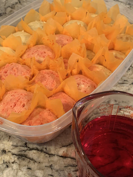 Image shows a rectangular shaped plastic container filled with orange lined white cupcakes. The ones closest to the front are pink with the ones furthest away white. The jug, at the front, is filled with 3 cups of pink/red liquid. 