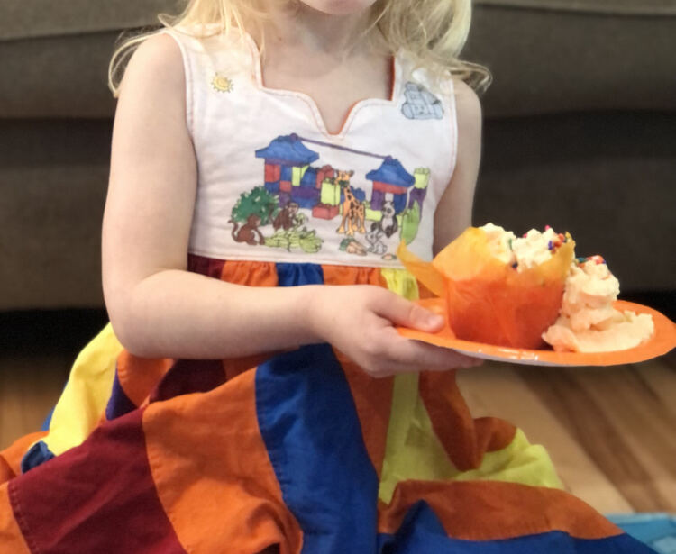 Image shows Zoey in a peppermint swirl dress holding an orange plate with a slightly blurred orange cupcake with orange whip cream and rainbow sprinkles on top. 
