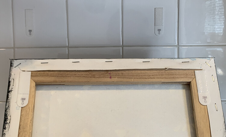 Image shows the back of the painting with the two command strips attached vertically to either corner. Behind it you can see the other two matching strips attached vertically to the white tiles. 