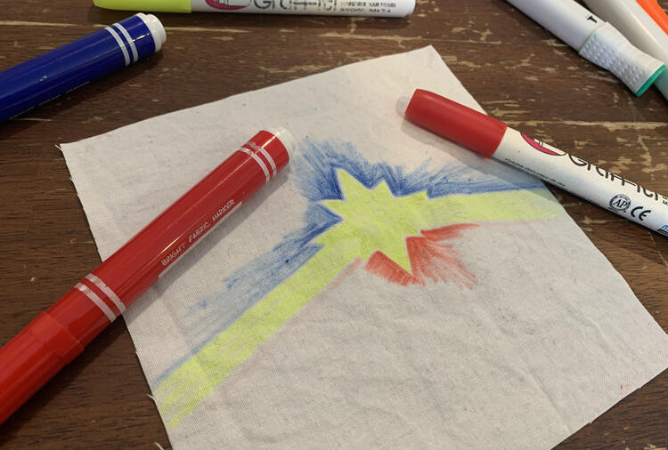 Image shows an upside down Captain Marvel logo on a square of white woven fabric. There are two red fabric markers laid over top. 