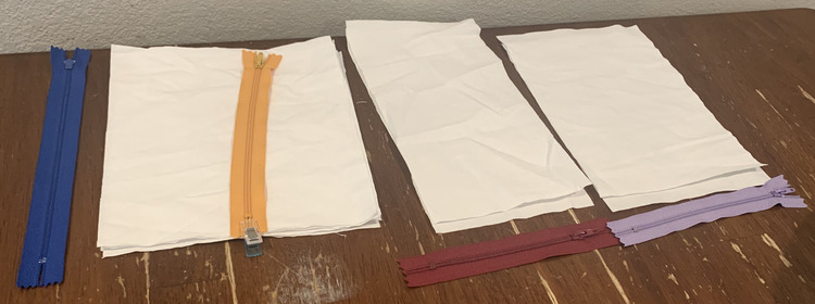 Image shows four zippers with three piles of rectangular white fabric. On the left the blue zipper is alone (waiting for orange fabric to be cut) while the next zipper is clipped to a pile of four rectangular fabric. Beside it there are two piles of two rectangles of fabric with a matching zipper beside each one. 