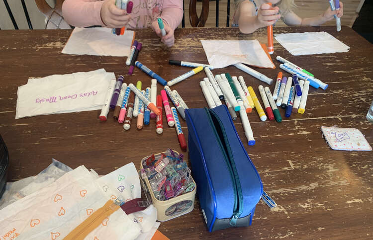 Image shows the kids grabbing fabric markers from a pile in the center while white rectangles of fabric sit in front of them. Closer to the bottom sits a pile of clips, and empty pouch, and a stack of fabric and zippers. 