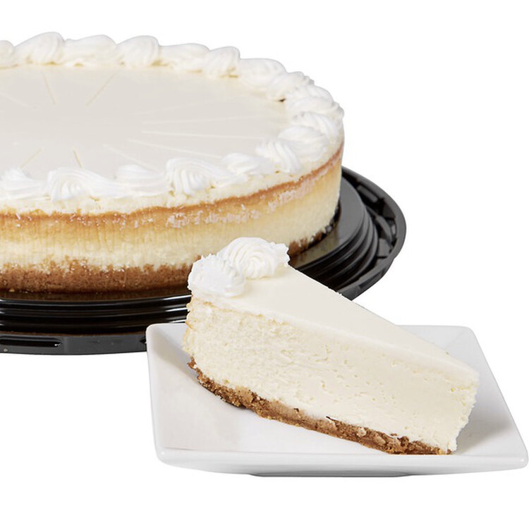 Image shows a whole cheesecake on the black plastic base without the clean lid overtop. The left side is cut off and in front of it, on a white square plate, sits an unadorned slice of cheesecake. 