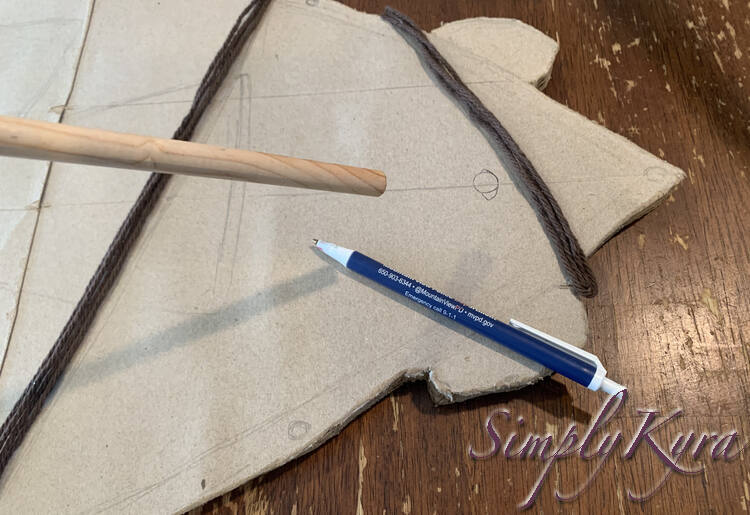 Image shows the front of the skateboard with the dowel held above it, a circle marked near the brown yarn, and the pen set to the side. 