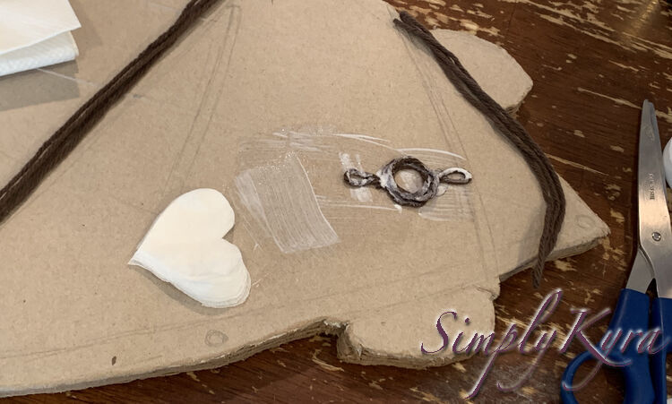 Image shows the brown circle with a loop coming from either end glued down to the cardboard skateboard and coated in more white. The section underneath has more glue showing where the heart napkin will go next. 