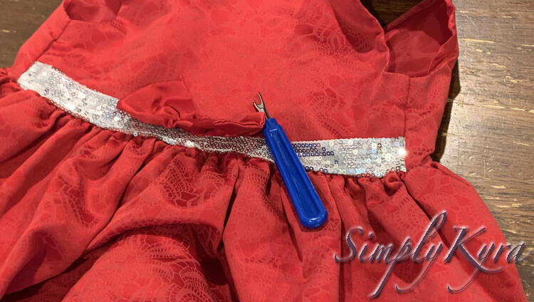 Image shows the center half of a fancy red dress with a white and silver sequined section along the front of the waist. On the center is a mostly removed red bow with a blue handled seam ripper laid out beside it.  