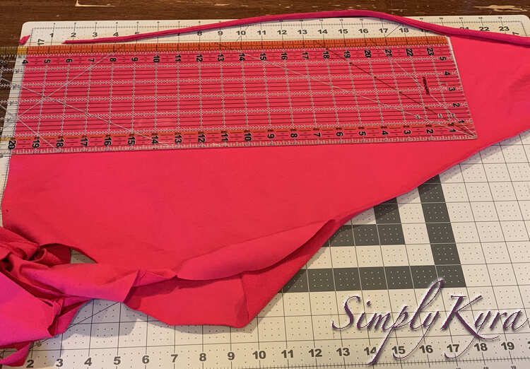 Image show a large triangle of pink knit fabric with a quilting ruler laid over the edge. That edge is trimmed off and the end is cut making a twenty inch long section from the remnant. 