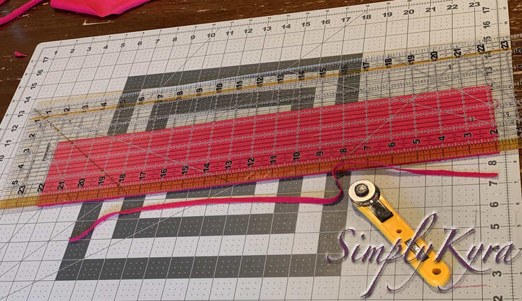 Image shows the pink strip measuring twenty by 3.25 inches now. The excess thin strip is cut off the edge with a yellow rotary blade off to the side. 