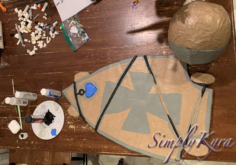 Image is an overview of the helmet and visor laid out on the kitchen table. All the grey is painted, the blue gem on the skateboard, and most of the necklace. To the left you can see the black bottle of paint upside down on the paint dish to get it all out while the other paints are scattered around. In the back you can see some of the LEGO my husband is building as we sit together at the table.  