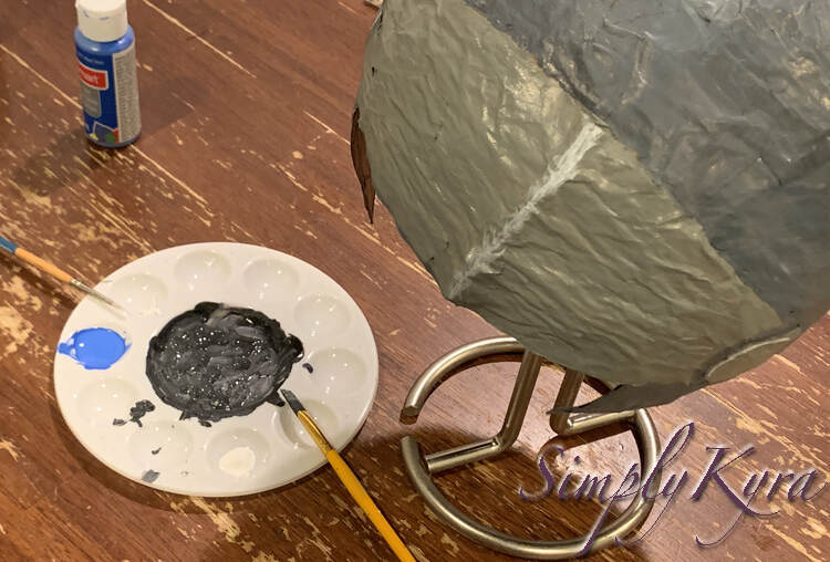 Image is taken from above looking down at the helmet, to the right, on the paper towel holder, and the paint plate and paints to the left. At this point the helmet is fully painted although the visor still needs the openings added. 