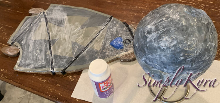 Image shows the helmet resting on the paper towel holder over a sheet of paper towel. In the background sits the Mod Podge and skateboard. Both items are coated in streaks of white Mod Podge showing where it's still wet. 