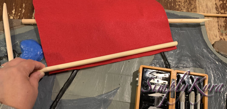 Image shows me laying the second dowel up along the bottom of the horizontal felt. Below it sits the rest of the X-Acto knives while underneath it lays the skateboard and extra pieces. 