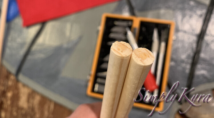 Image shows the two dowels held up with the ends cut cleanly. In the background you can see a blurred knife set, skateboard, table, felt, and dowels. 