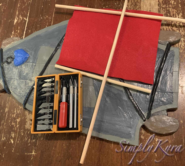 Image shows the sail laid out with the three mast pieces so it looks almost complete. Underneath it lays the skateboard and my table while beside it sits the open X-Acto knife container. 