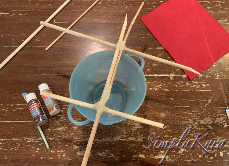 Image shows the unpainted dowel and tape mast laid out on the top of the blue bowl with the paints, paintbrush, extra doweling, and red felt in the background around it. 