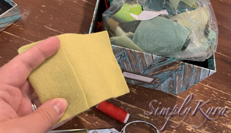 Image shows my left hand holding a rectangle of felt with the end folded over. Behind it you can see some red thread and a photo box filled with felt. 