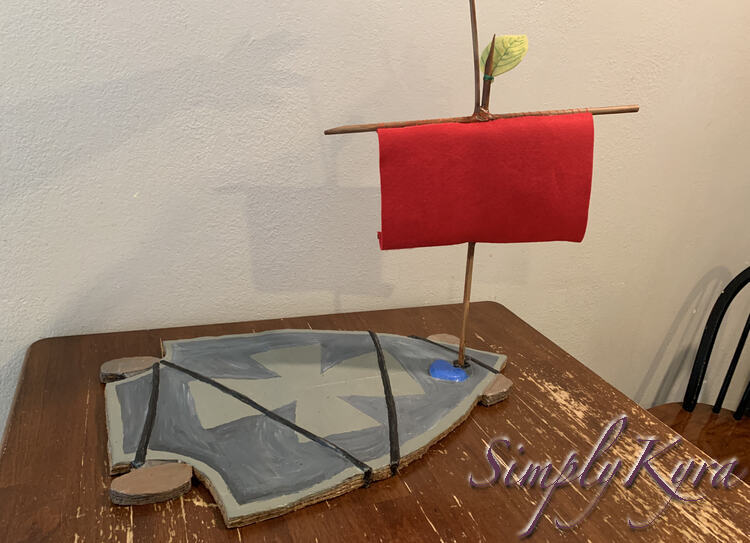 Image has the sail upright in the skateboard sitting on the table. You can see a shadowy arm against the wall holding the top of the mast in place