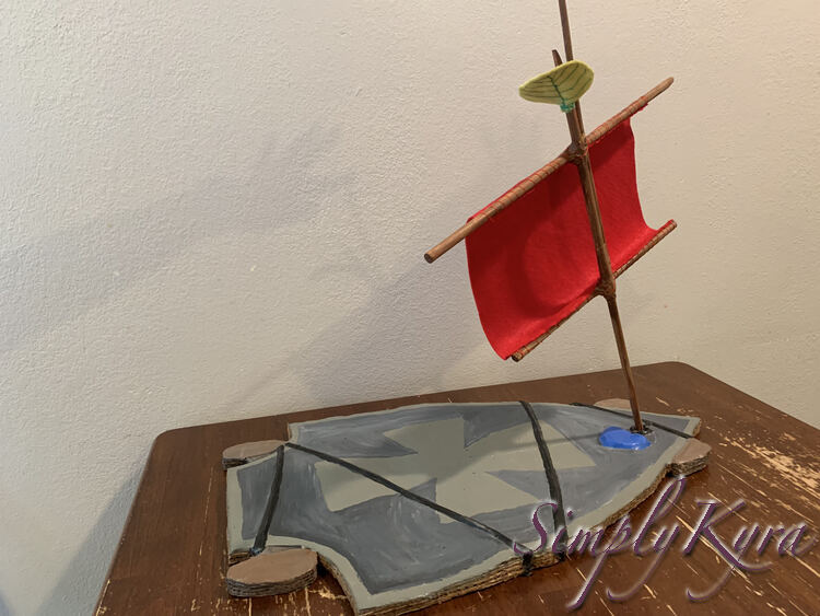 Image has the sail at an angle as if it had to catch the wind. The base is resting in the skateboard and a shadowy arm can be seen against the wall holding the top of the mast in place.