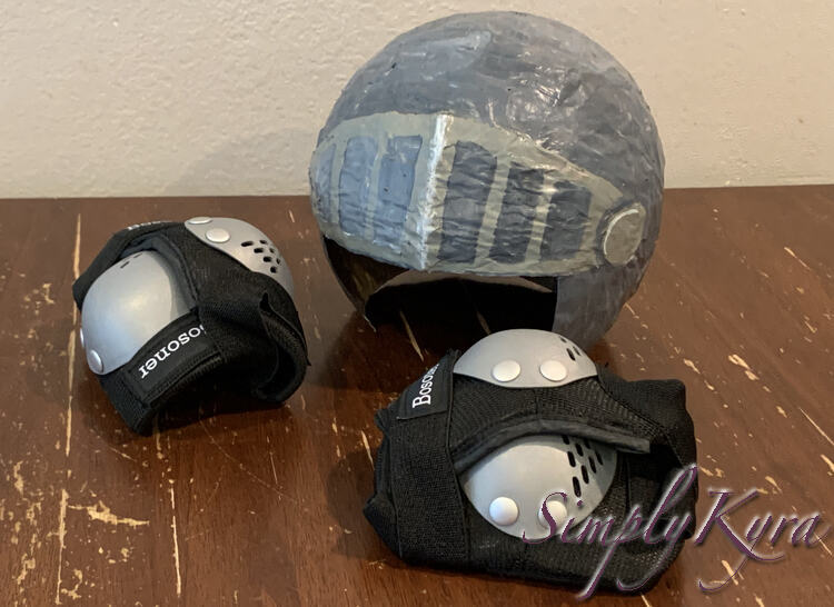 Image shows the helmet resting on the table. Beside it are two elbow pads attached together while in front of it are two kneed pads together. 