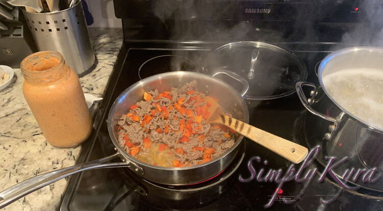 Image shows a skillet with ground beef, red bell pepper, grease, and a wooden flipper. To the right is a boiling pot of pasta in water and to the left is an opened canning jar of leftover soup.