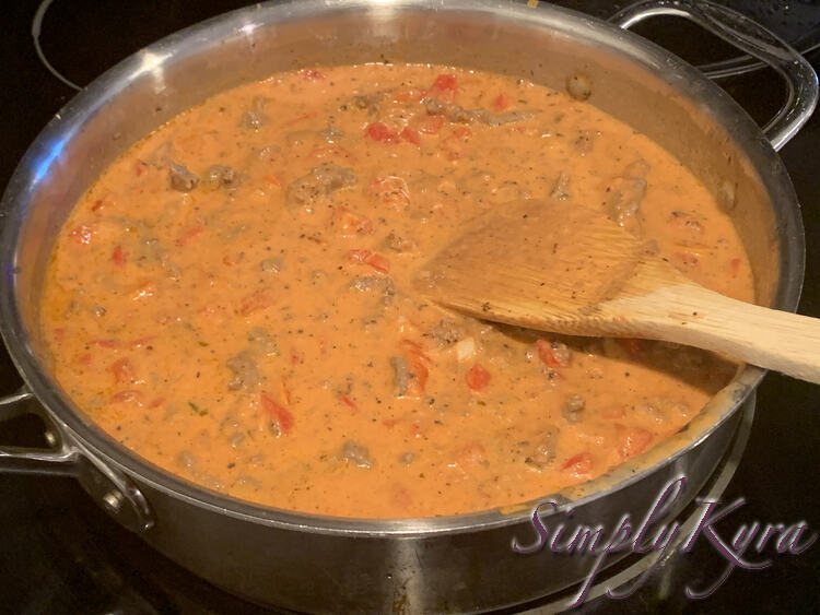 Image shows a closeup of the skillet with the wooden spatula laid in it. The sauce is orangey-red with floating bits of ground beef, red pepper, and spices in it. 