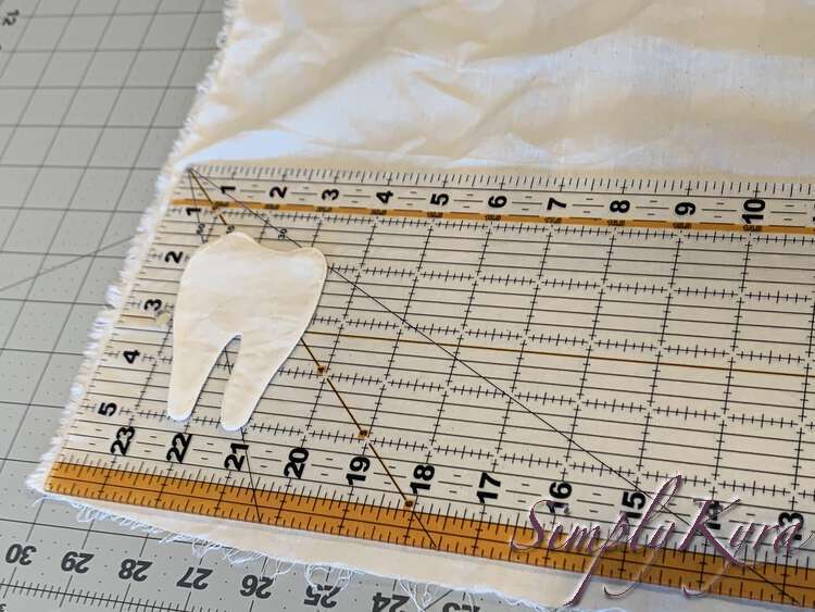 Image shows white fabric laid out over a cutting mat. In the 90 degree raw edged corner is laid out the clear quilting ruler and above that the white tooth. 