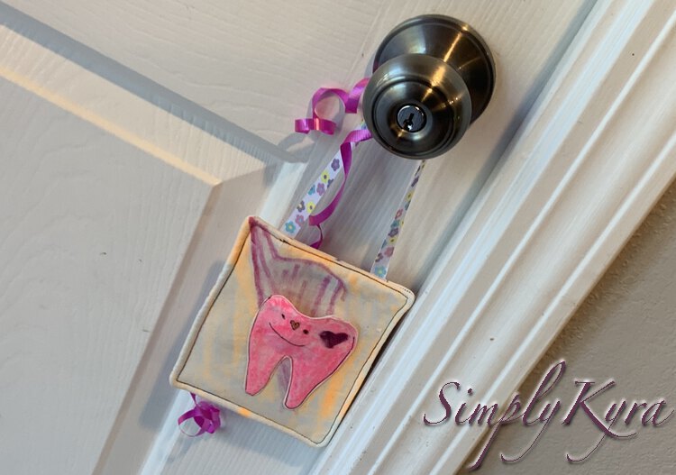 Image shows Ada's final tooth fairy pouch hanging from a silver doorknob in front of a white door with a pink curling ribbon behind it. The tooth itself now has a heart on it's one cheek.  