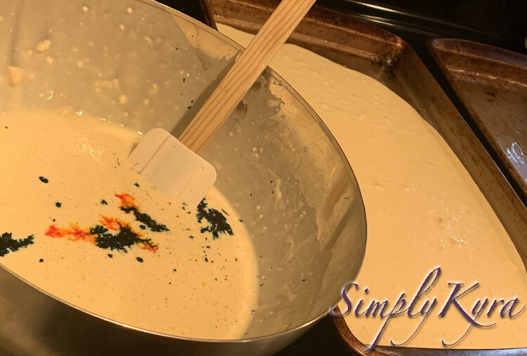 Image shows a bowl of white pancake batter with splotches of yellow (kind or orange looking) and blue. A white spatula sits in the bowl. Off to the right you can see one mostly full sheet pan and one empty one beside it.