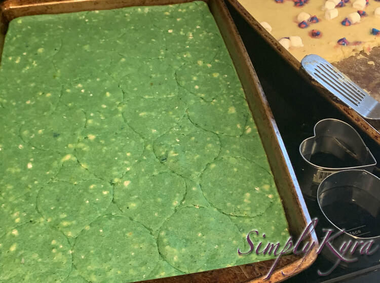 Image shows a pan of green pancake with hearts and the odd circle cut into it. Beside it sits two metal cookie cutters, one circular and one heart shaped, and then the half filled pan of s'more inspired pancakes and a metal flipper for dishing. 