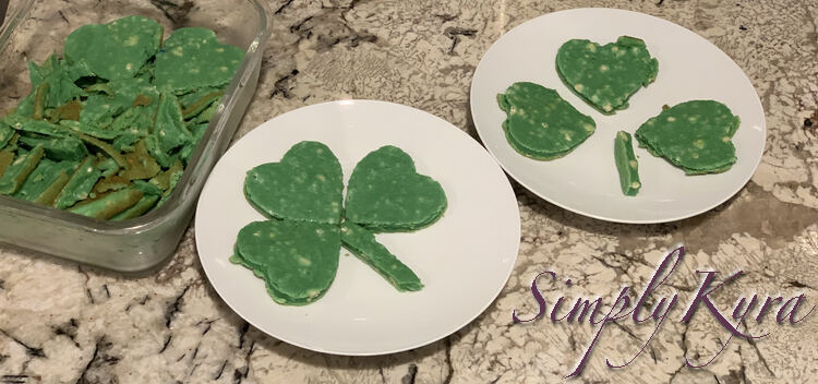 Image shows a partially assembled shamrock on the right plate, and fully assembled one on the left plate, and a container of leftover green pancake pieces on the far left. 