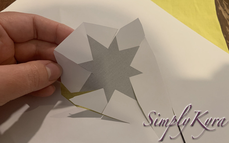 Image shows my hand holding up a piece of paper with the grey star printed on it. Each point has one edge cut while the opposing point is holding the paper in place. Behind it sits the rest of the paper and the bright yellow fabric I'm going to use for the star. 
