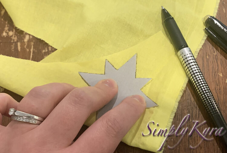 Image shows me holding the grey paper star in place on the yellow fabric. A black disappearing ink pen sits beside. You can see an outline around the outside of the star were I already traced around it. 
