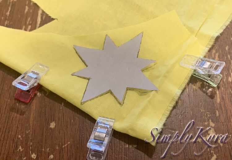 Image shows the yellow fabric folded over with the grey paper star placed lightly on top with the outline already marked. Three clips keep the fabric in place. 
