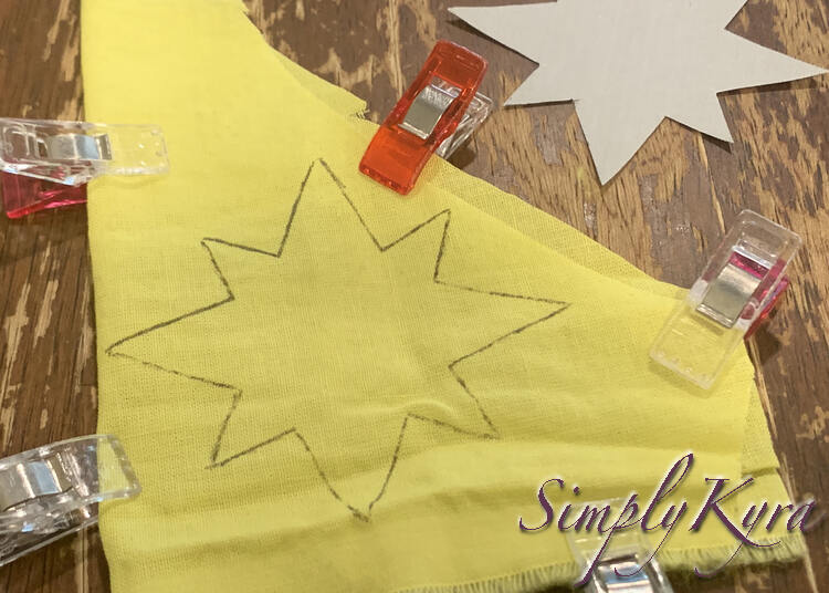 Image shows a closeup of the yellow fabric with the black inked outline of a star. There are five sewing clips placed around the fabric and the paper template is off to the side at the top of the photo. 