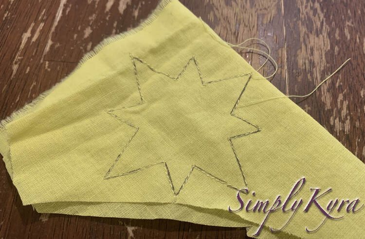 Image shows the same scrap of fabric on the kitchen table. The black star's outline is obscured by yellow thread overlapping it. The loose thread ends show where the stitching was started and ended. 
