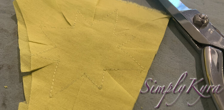 Image shows the same fabric with the yellow star but now there are cuts leading to each inner corner in the star and the metal scissors lay beside. 