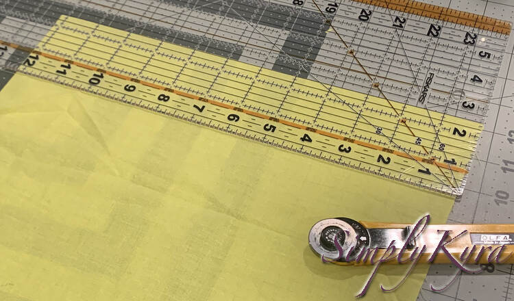 Image shows a rectangle of yellow fabric on a cutting board. The clear ruler overtop shows I'm about to cut a strip of fabric thats 2.5 inches by 12. A rotary cutter sits near the bottom. 