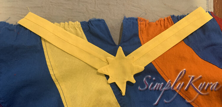 Image shows the star laid out below the slit on the blue fabric. The strips are angled up going to the top of the dress making a triangle. Behind the yellow fabric you can see the yellow, orange, blue, and red swirls. 