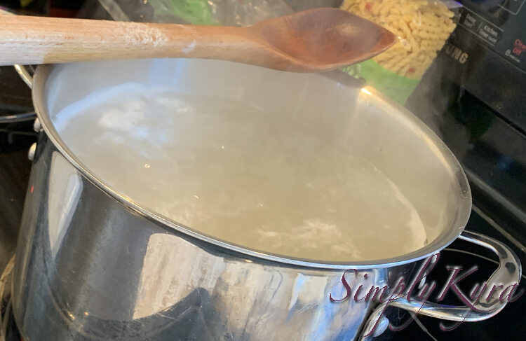 Image shows a pot of steaming water with the haze of pasta already in it. A wooden spoon is laid on top and a you can see the empty pasta bag beside the second bag of pasta in the back. 