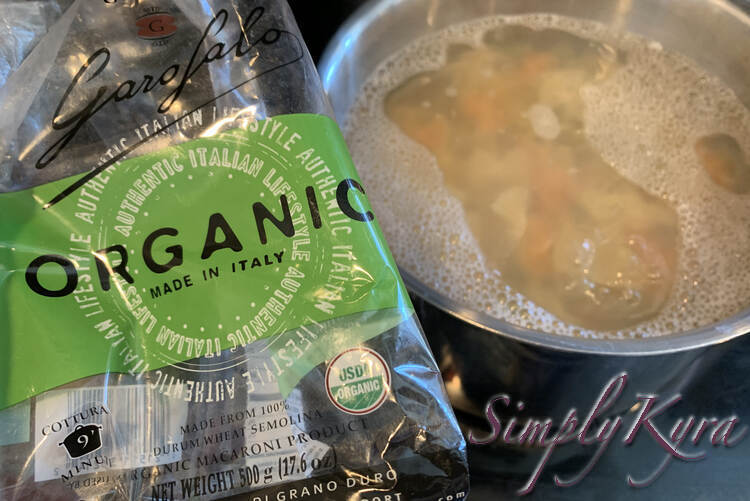 Image shows the empty second bag of pasta held up showing the pot containing both packages of pasta and boiling water in the background. 