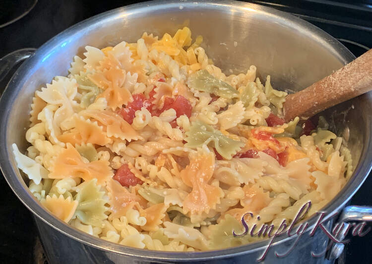 Image looks down at a pot filled with beige gemilli pasta and tricolored farfelle. The three colors are mixed with brighter orange cheese and red tomatoes. 