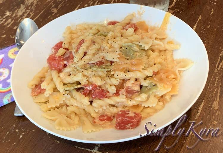 Image shows a mix of beige, red, orange, and green pasta mixture on a while plate sprinkled with black flecks. A metal spoon and purple rainbow and donut covered napkin sits beside. 