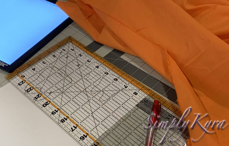 Image shows the printed teeth laid out on a cutting mat and obscured by a transparent quilting ruler. On top sits a red pen while orange fabric is laid out above and a turned on light box is to the left. 