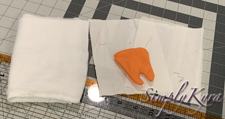 Image shows a folded piece of soft fabric with selvage at one end folded next to the slightly stacked two white rectangles of fabric and an orange puffed up tooth. Below them lays the cutting mat and quilting ruler. 