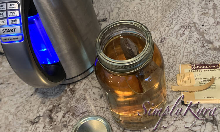Image shows the same canning jar filled with brown transparent liquid with a tea bag now floating at the top with a string attached to the outer edge under the lid. To the left is a electric kettle with light on and to the right is the empty tea packaging and tag. 